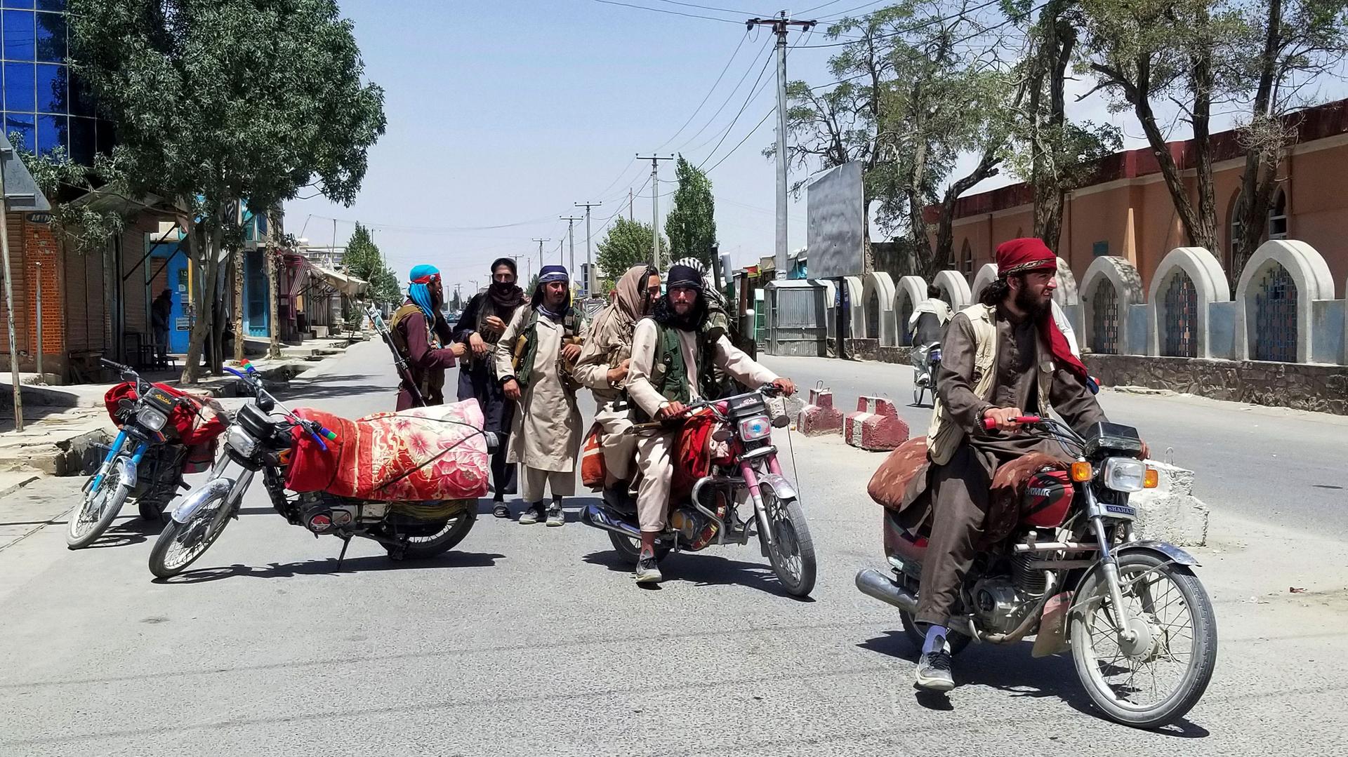 Several Taliban fighters are shown standing in a street or sitting on blanket-covered motorcycles.