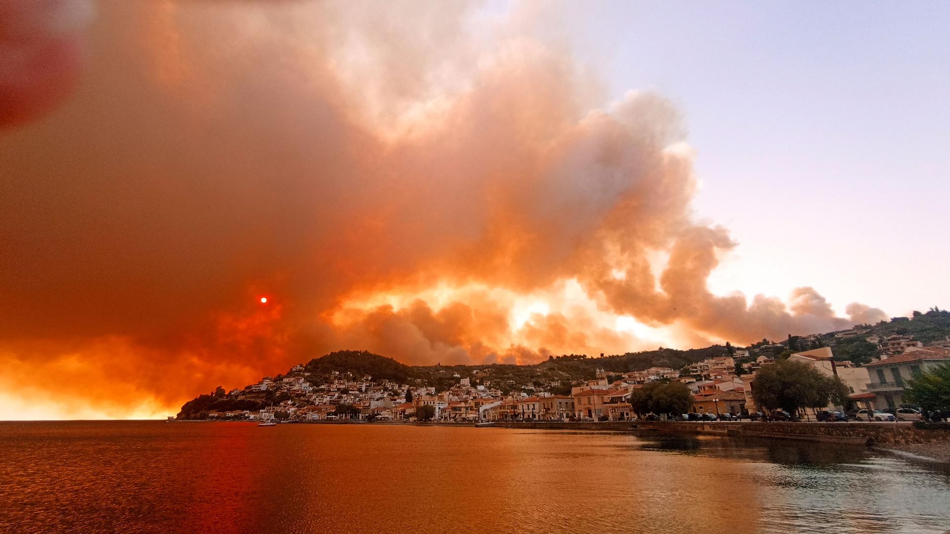 The shoreline of a village on the Greek island of Evia is shown with massive plumes of dark smoke rising in the distance.