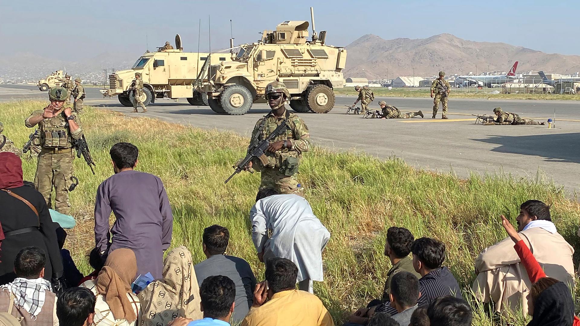 Several US military personnel are show standing guard next to the runways of the Kabul airport with two large military vehicles in the distance.