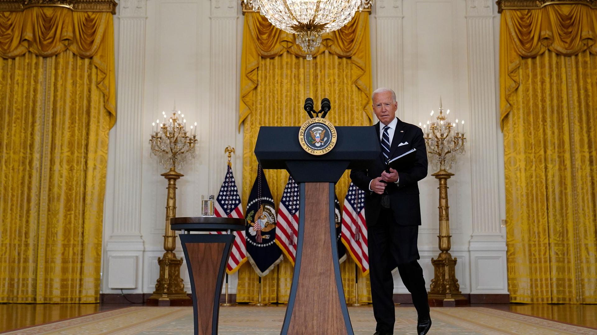 US President Joe Biden is shown walking toward a podium with the seal of the US presidency and holding a folder.