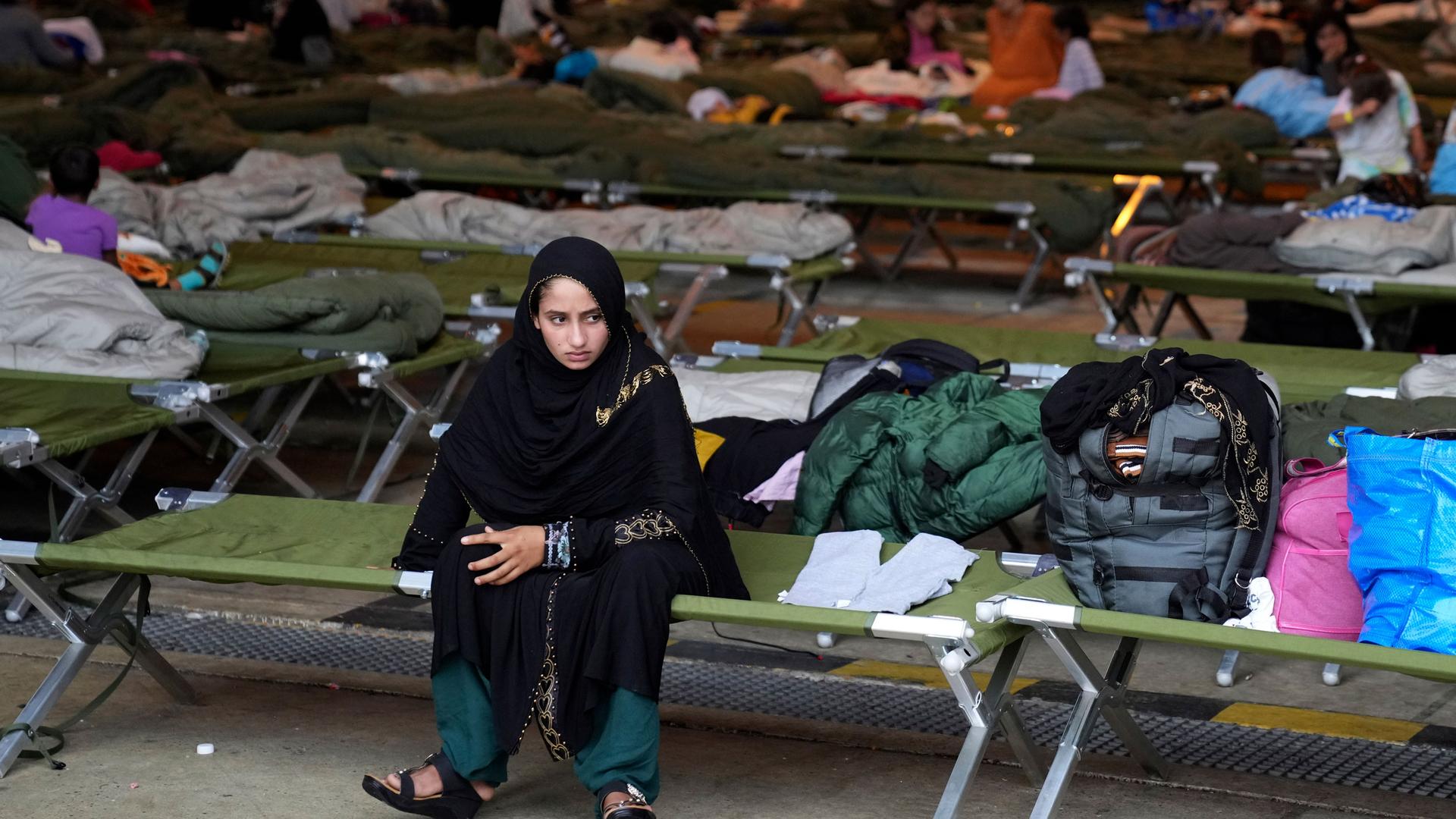 A young woman is shown sitting on a green cot, among dozens over other cots, each with personal belongings on top.