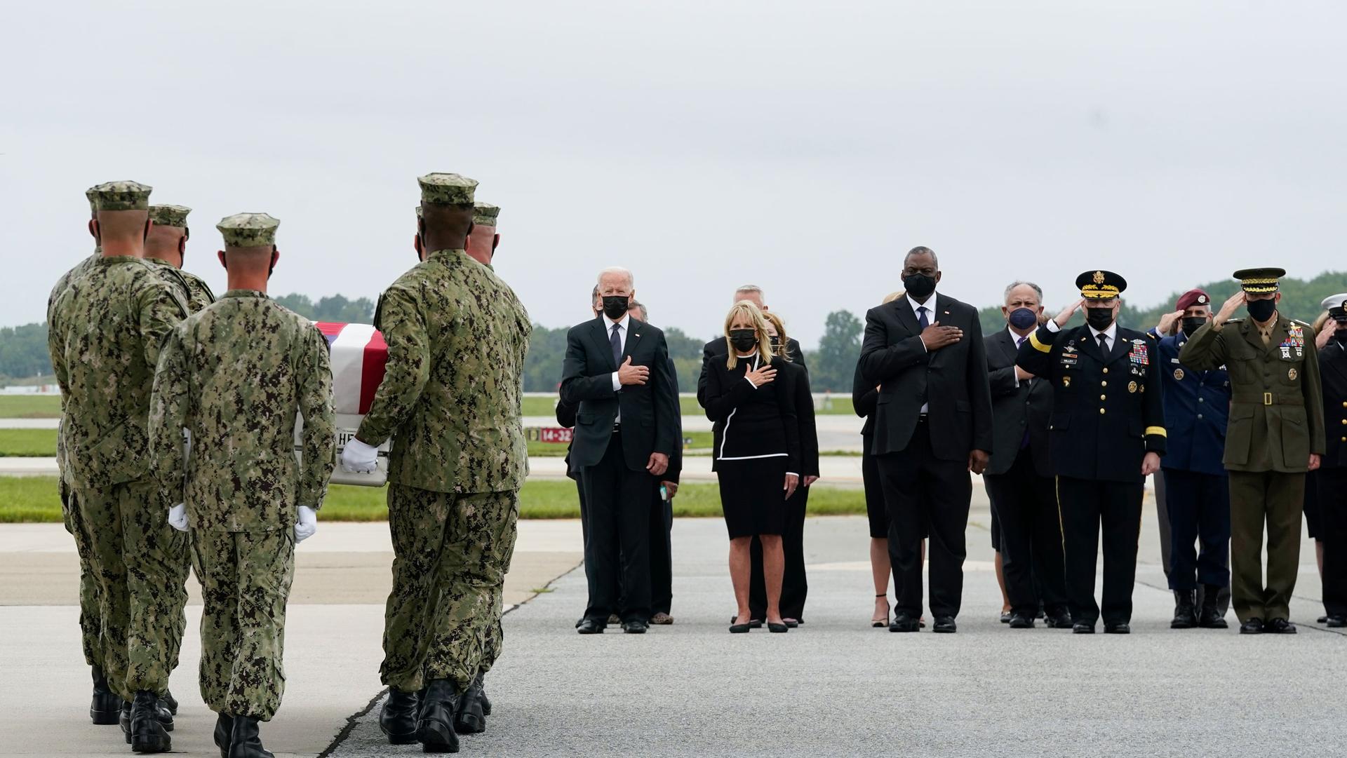 A group of military personel are shown carrying a flag-drapped coffin and walking toward US President Joe Biden and other cabinet officials.