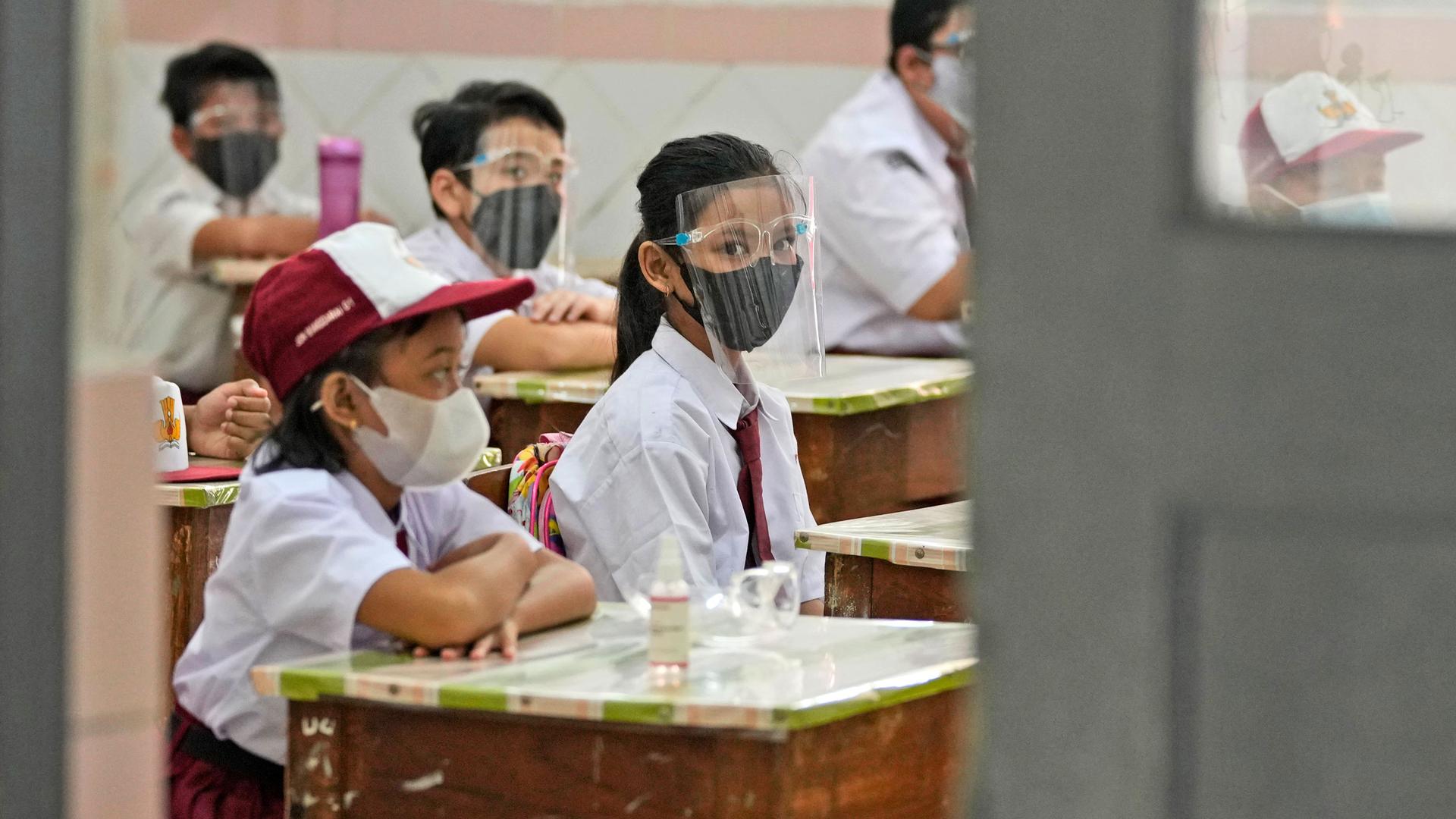 Several young school children are shown sitting at desks with one girl looking back at the camera while wearing a clear face shield and mask.