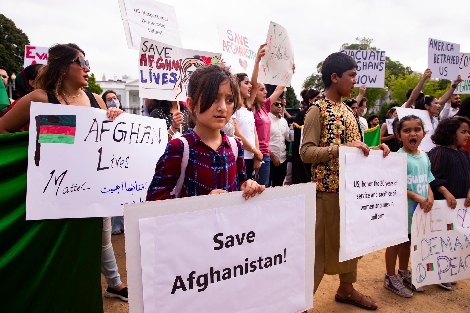 Women holding signs saying "save Afghan lives" and "Afghan Lives Matter" at a protest, with the White House in the background