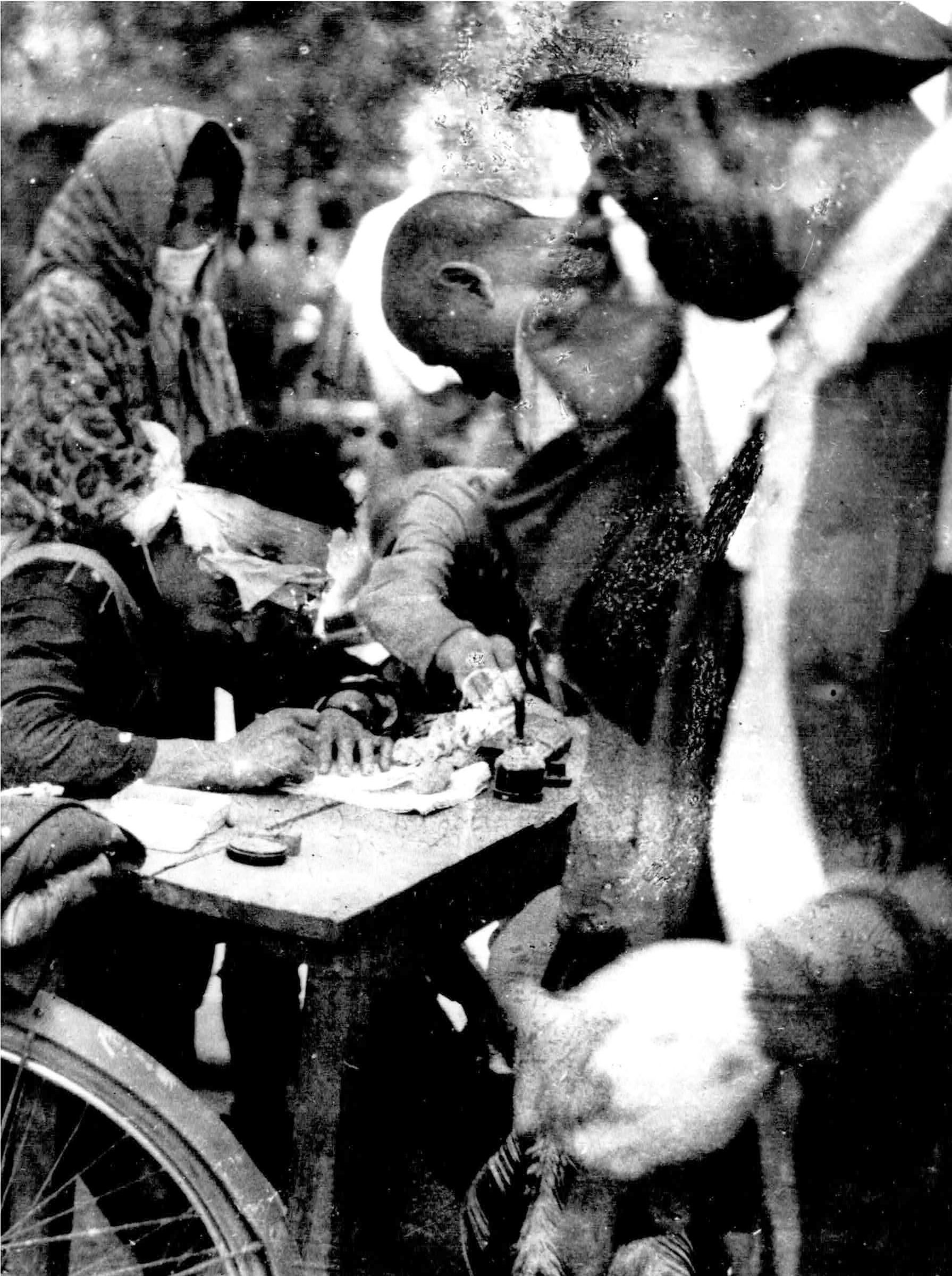 A police officer is shown sitting at a table while wearing a bandage on his head and writing on paper with several people standing around him.