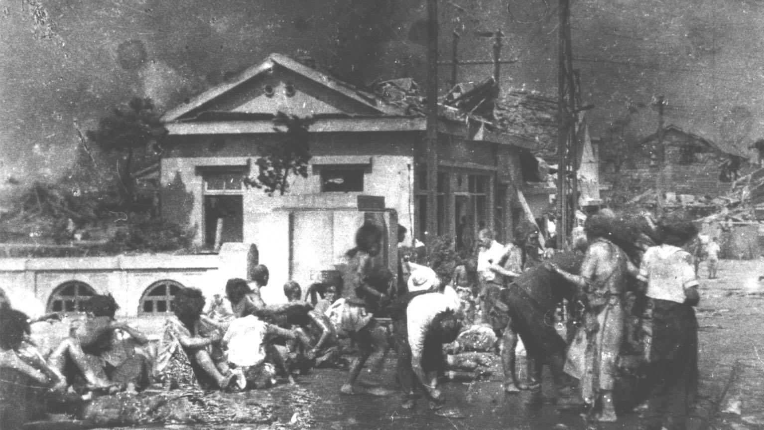 A crowd of survivors are shown standing and sitting on a bridge with several damaged buildings in the distance.