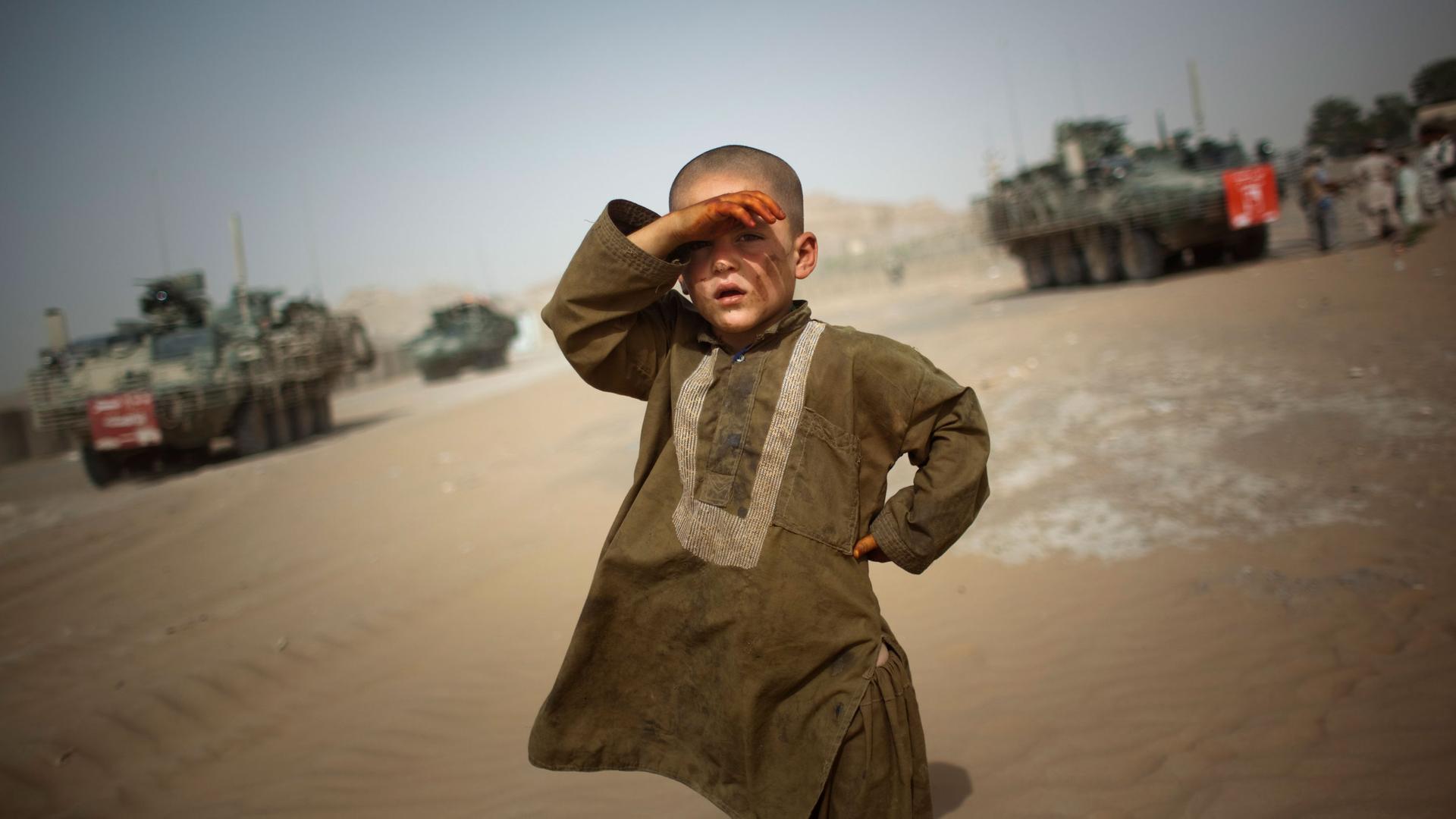 A young boy is shown with his right hand up to his face shielding the sun with armored military vehicles shown in the distance.