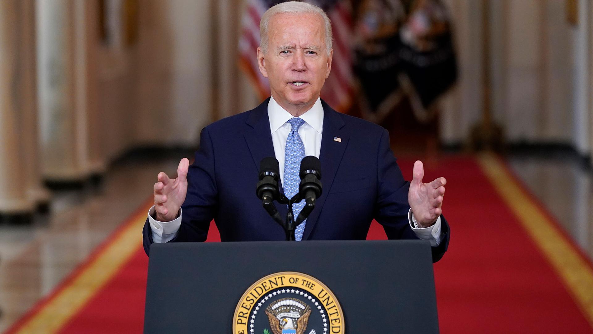 US President Joe Biden is shown standing at a podium and speaking into a microphone with his hands raised slightly.