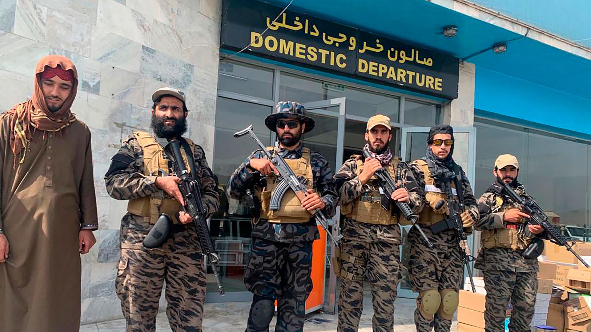A group of six men are shown holding guns and wearing military fatigues with the domestic departure entrance to the Kabul airport.