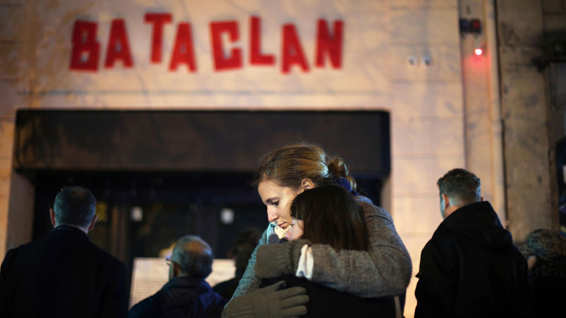 Two women are shown hugging each other with the entrance and red-lettered sign of the Bataclan concert hall in the background.