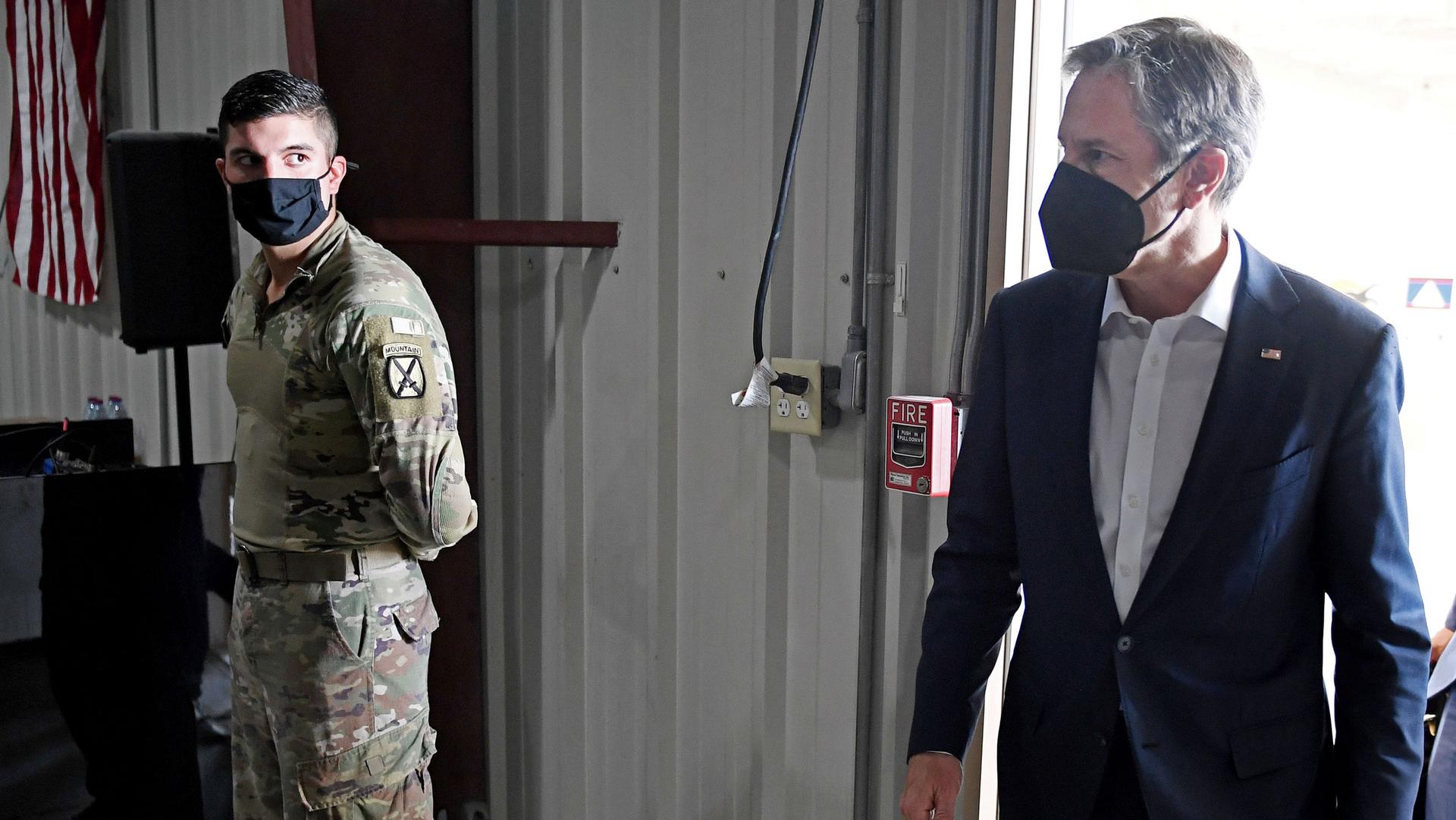 US Secretary of State Antony Blinken is shown looking to his right while walking and wearing a dark face mask with a solider stands in the distance.