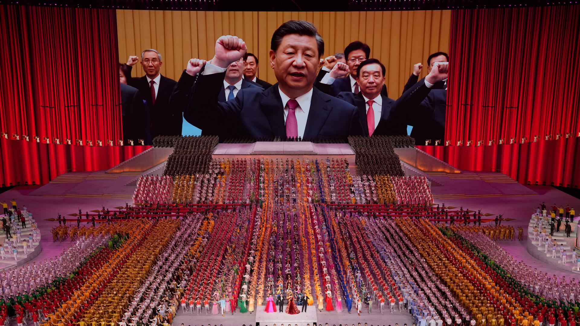 A large auditorium is shown filled with people in color-coordinated clothing with Chinese President Xi Jinping projected on a large screen in the distance.