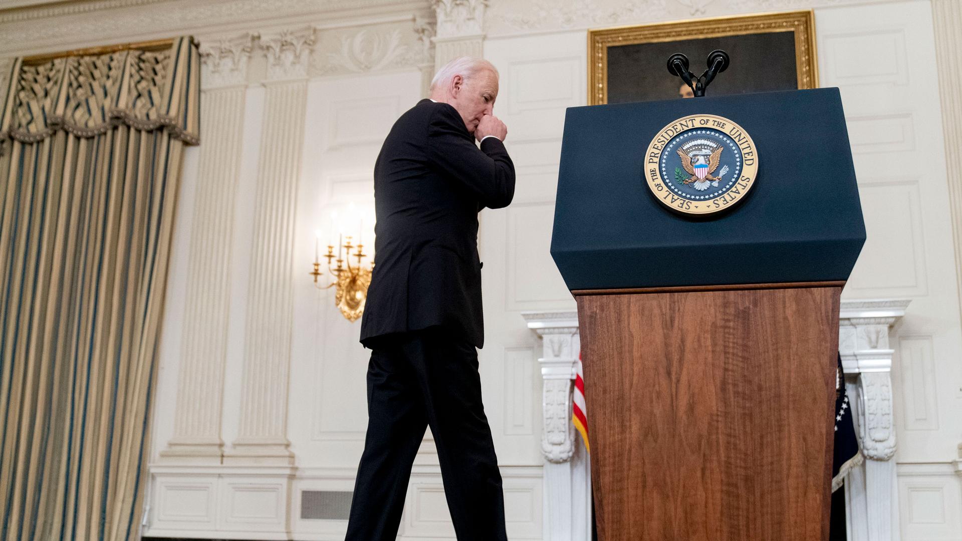 US President Joe Biden is shown wearing a dark suit and walking toward a podium with the President of the United States seal on it.
