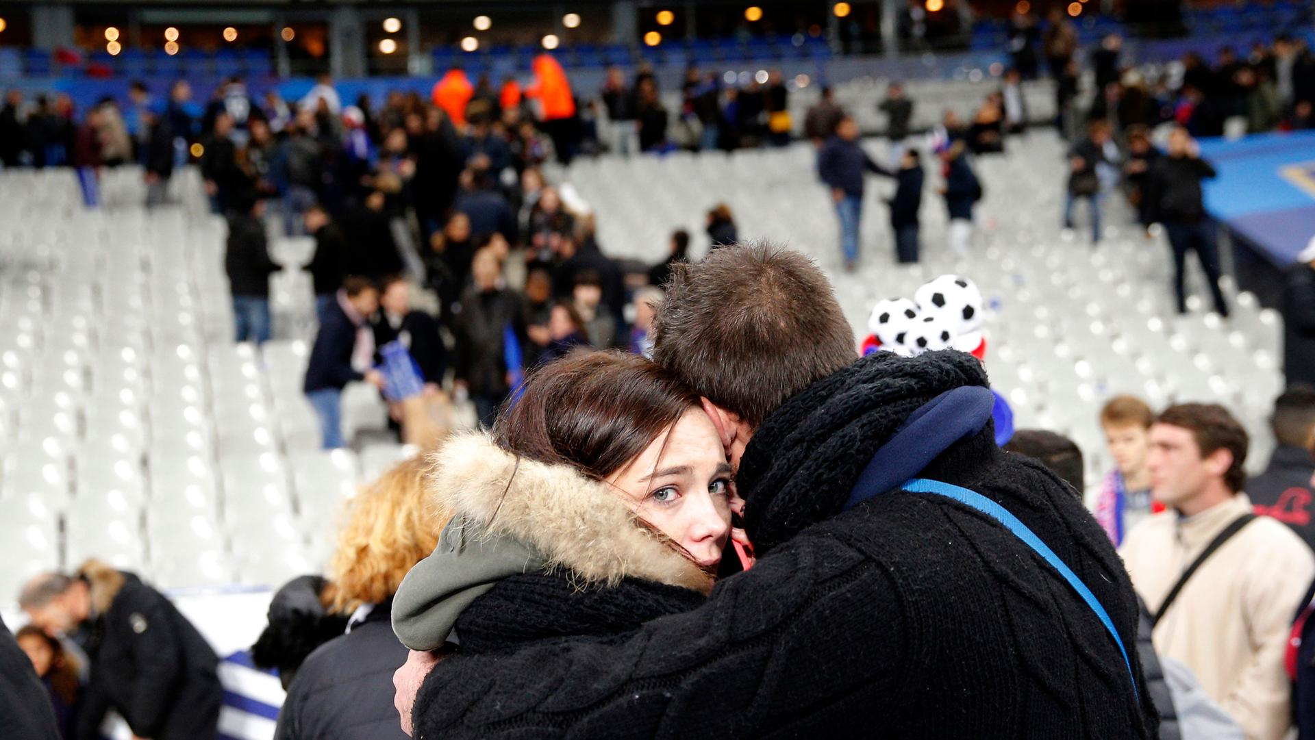 A man is shown hugging a woman who is looking into the camera with empty stadium seats in the distance.