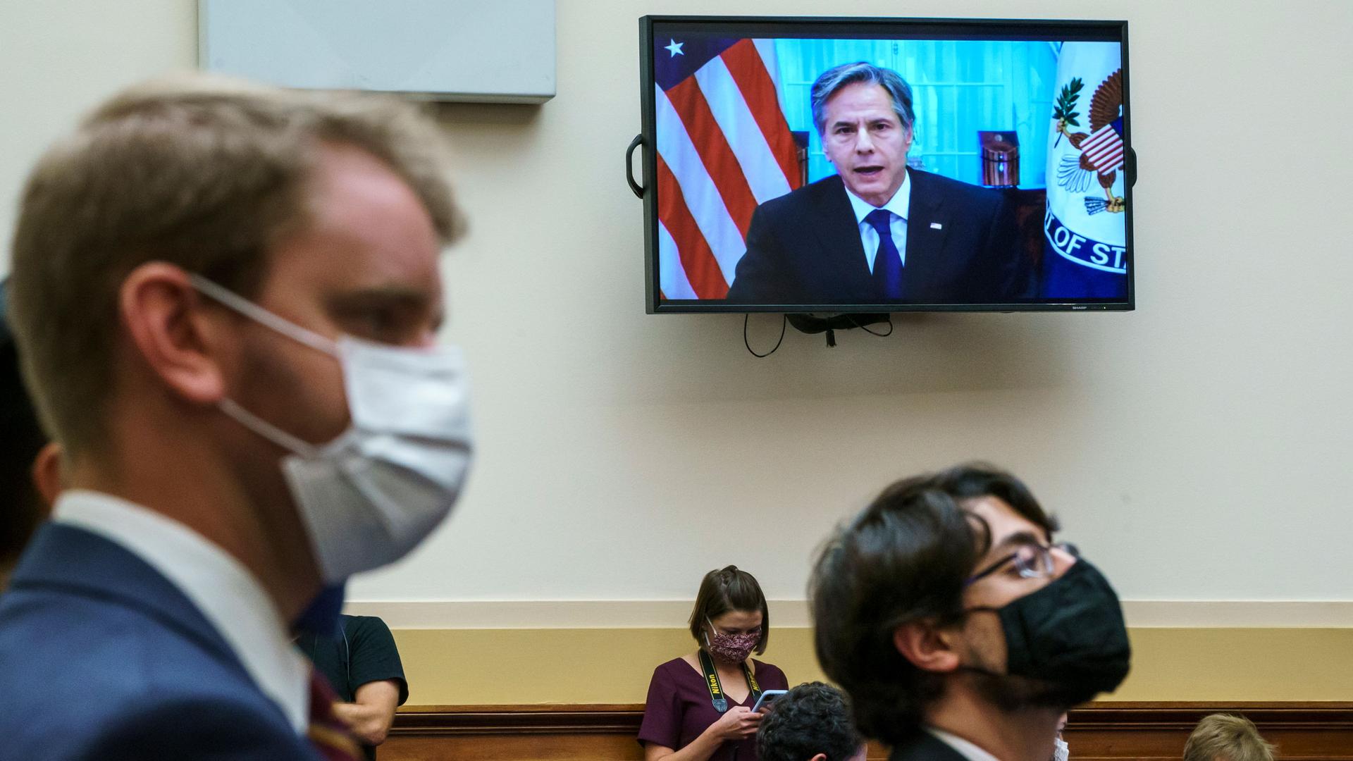 US Secretary of State Antony Blinken is shown on a TV hanging on the wall in the distance with several people wearing face masks stand in the nearground.