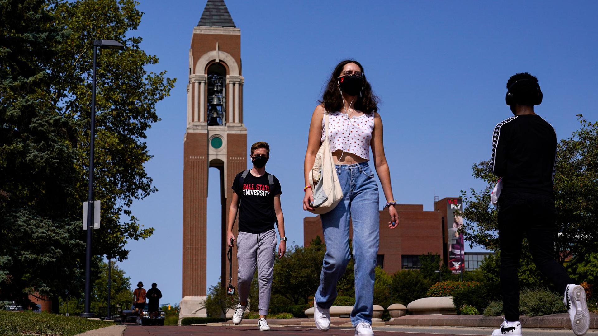Several people are shown wearing face masks and walking on a college campus with a ornate brick tower in the distance.
