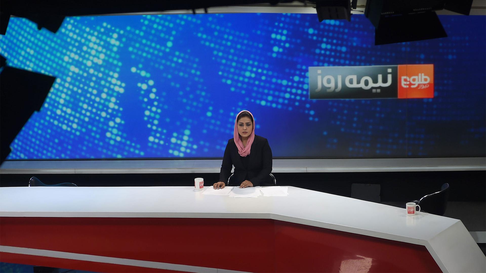 Woman news anchor wearing black outfit and pink headscarf looks into a camera in a Tolo News studio