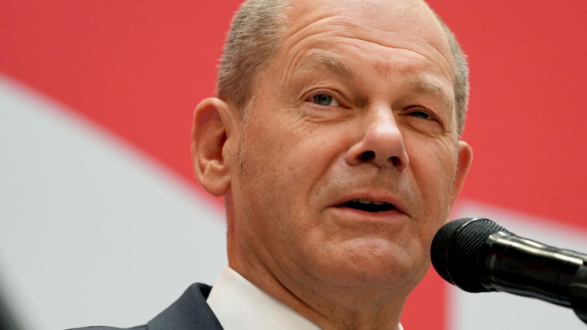 Olaf Scholz is shown in a close-up photoographer standing behind a microphone.