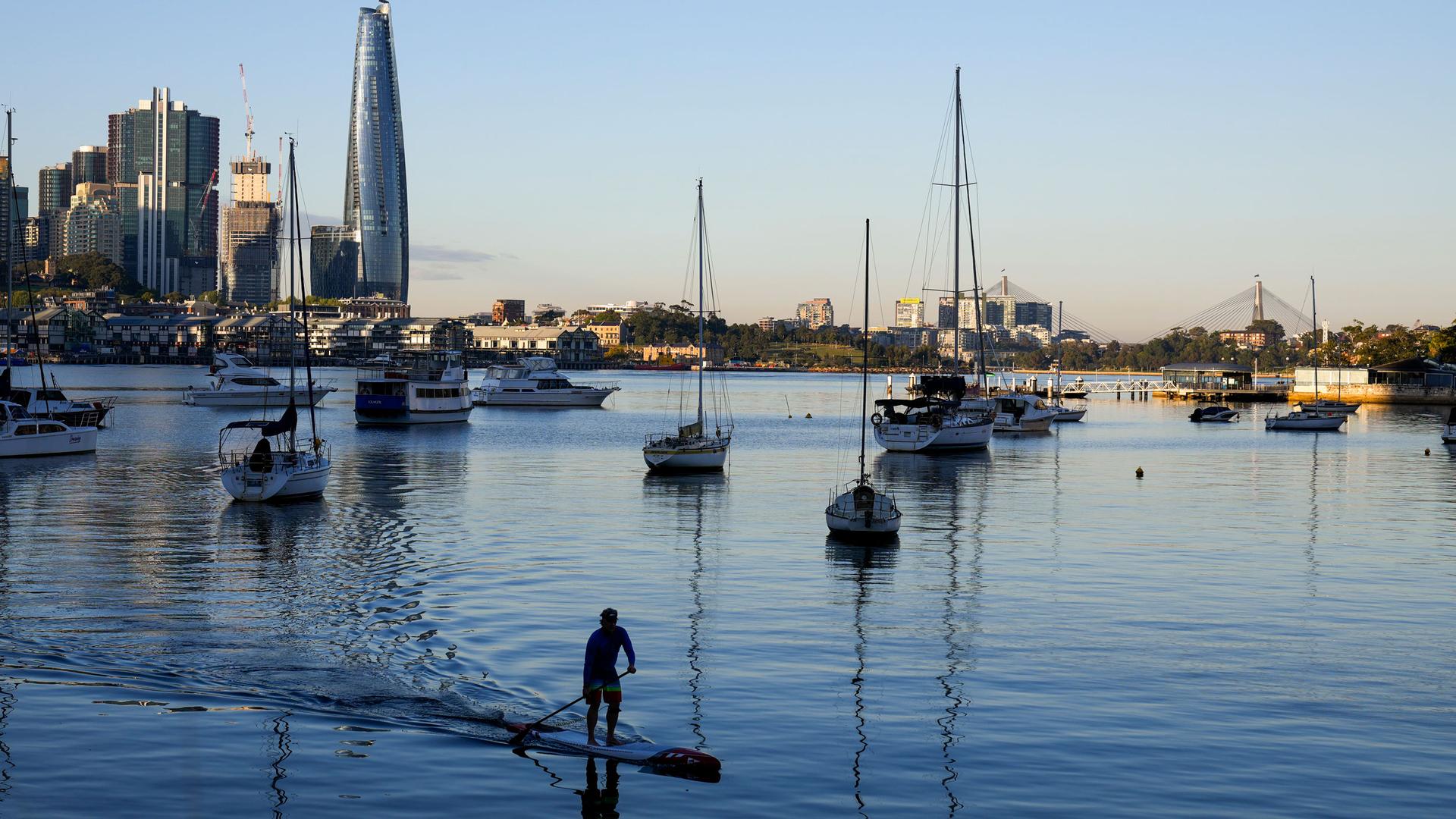 A man is shown on his stand up paddle board with several boats docked in the distance along with Sydney's skyline.
