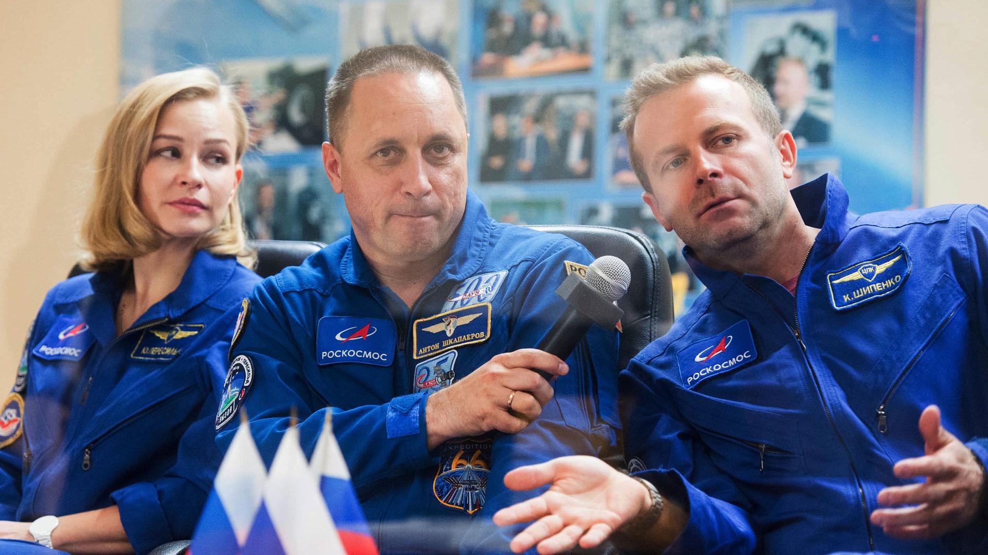 A woman and two men are sitting next to each other wearing blue Russian cosmonaut uniforms with the middle person holding a microphone.