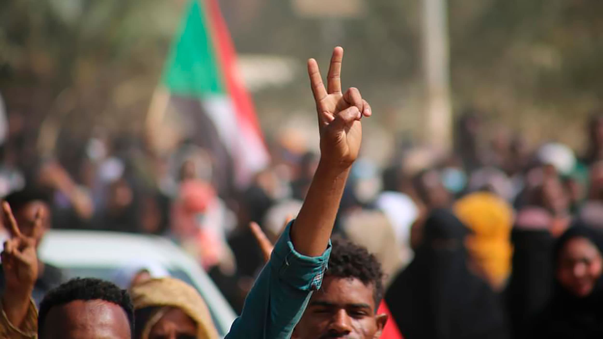 A protesters makes a peace sign with this fingers during a protest in Khartoum with the flag waving behind them.