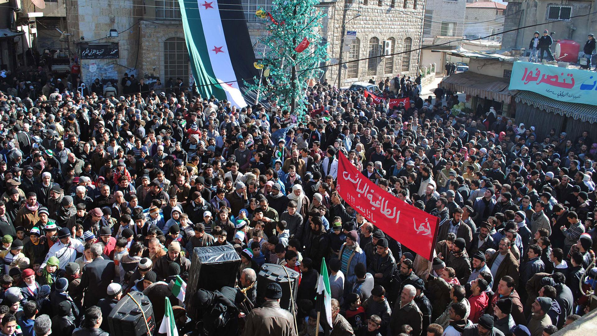 In this Friday Jan. 20, 2012, file photo, anti-Syrian regime protesters gather at a square as they hold an Arabic banner, center, reading, "Hey, the miserable, the tyrant, what else," during a demonstration at the mountain resort town of Zabadani, Syria.
