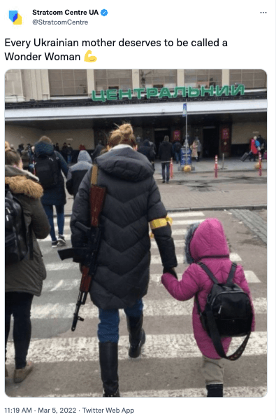 Screenshot of a tweet, showing Ukrainian woman crossing street holding hands with a young child, with an automatic rifle slung over her other shoulder