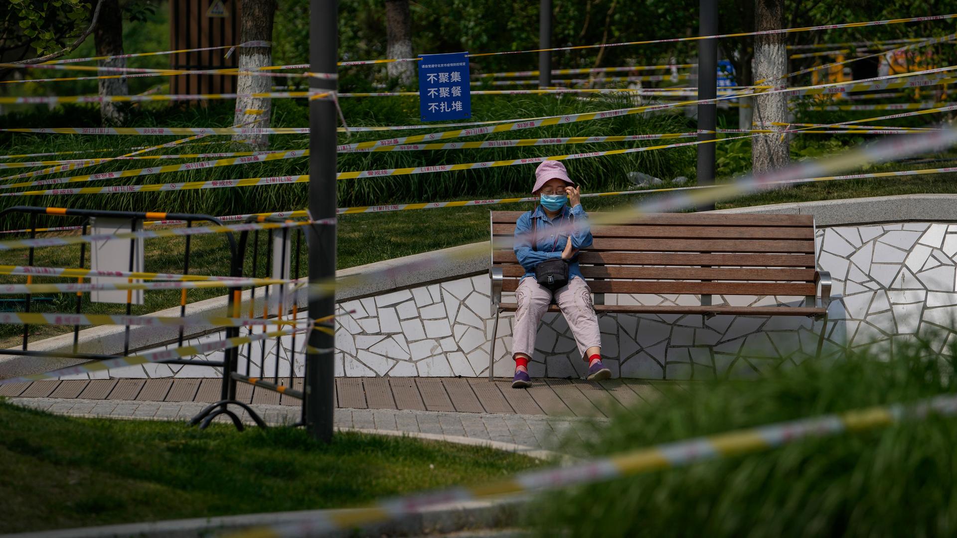 A woman wearing a face mask rests on a bench at the green space barricaded with fences and tapes to prevent residents from gathering due to pandemic measures on Thursday, May 19, 2022, in Beijing.