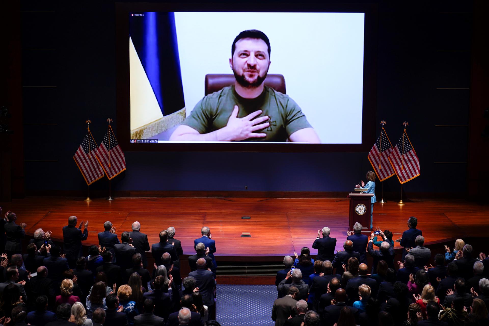 President Zelenskyy wearing a green t-shirt looks out of a large screen on the stage and puts his hand on his heart while the US Congress watches