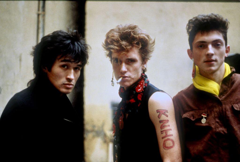 Three punk rockers standing a row, with tattoos and cigarettes