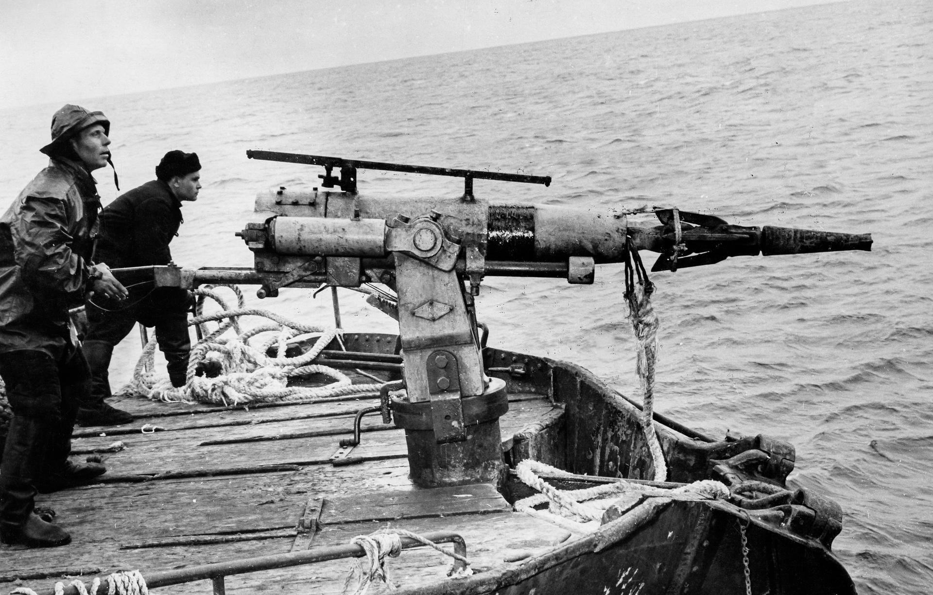 Black-and-white photo of two men in outdoor gear standing besides a large harpoon gun on a boat