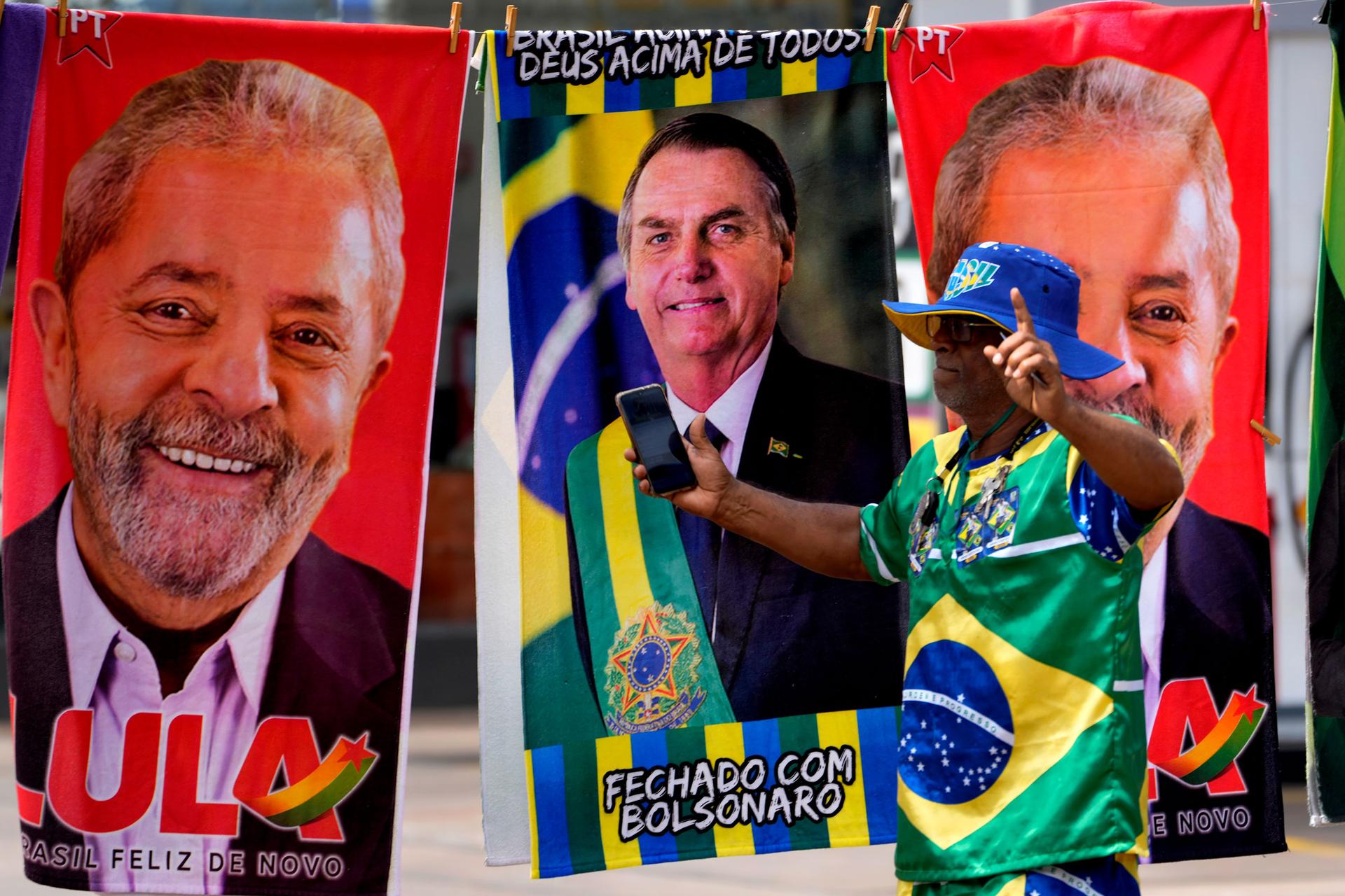 A demonstrator dressed in the colors of the Brazilian flag performs in front of a street vendor's towels for sale featuring Brazilian presidential candidates, current President Jair Bolsonaro, center, and former President Luiz Inacio Lula da Silva, in Bra