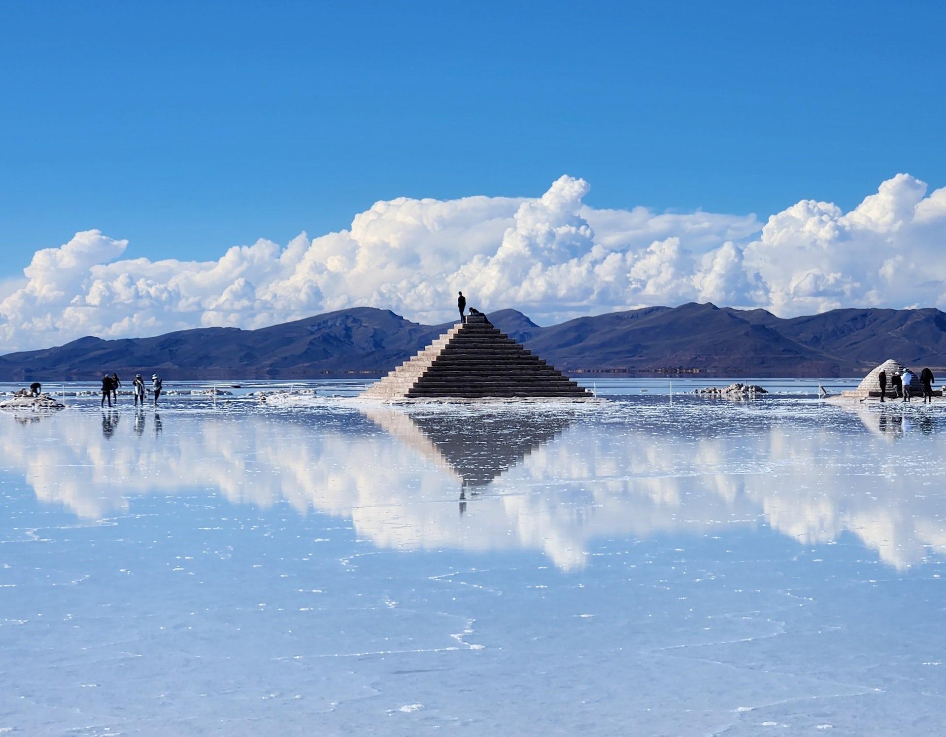 A person standing on a pyramid of dried salt, while the rest of the salt flat looks like the surface of a lake