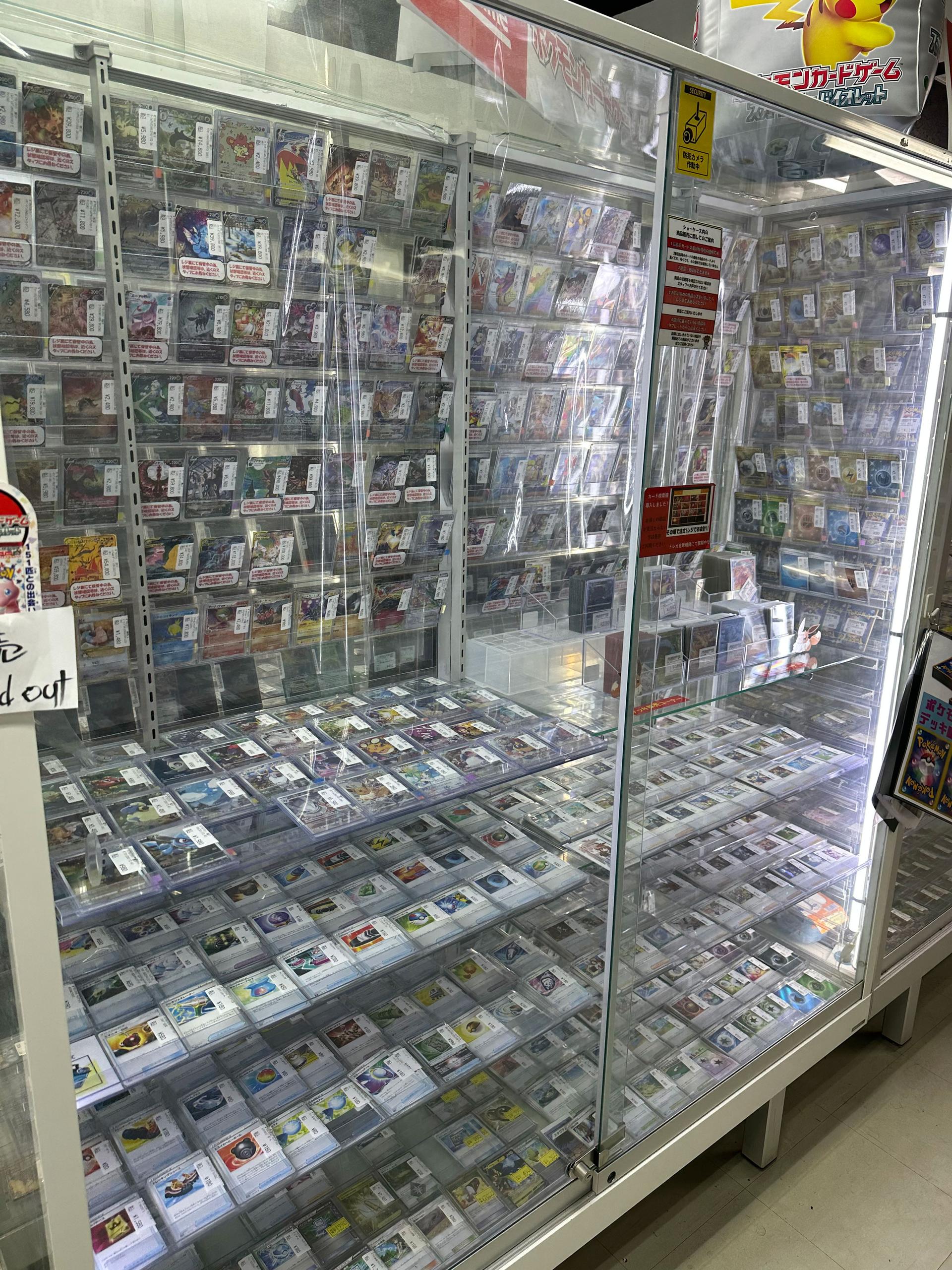 Pokémon cards are displayed in a glass cabinet. High-value cards are marked as 
