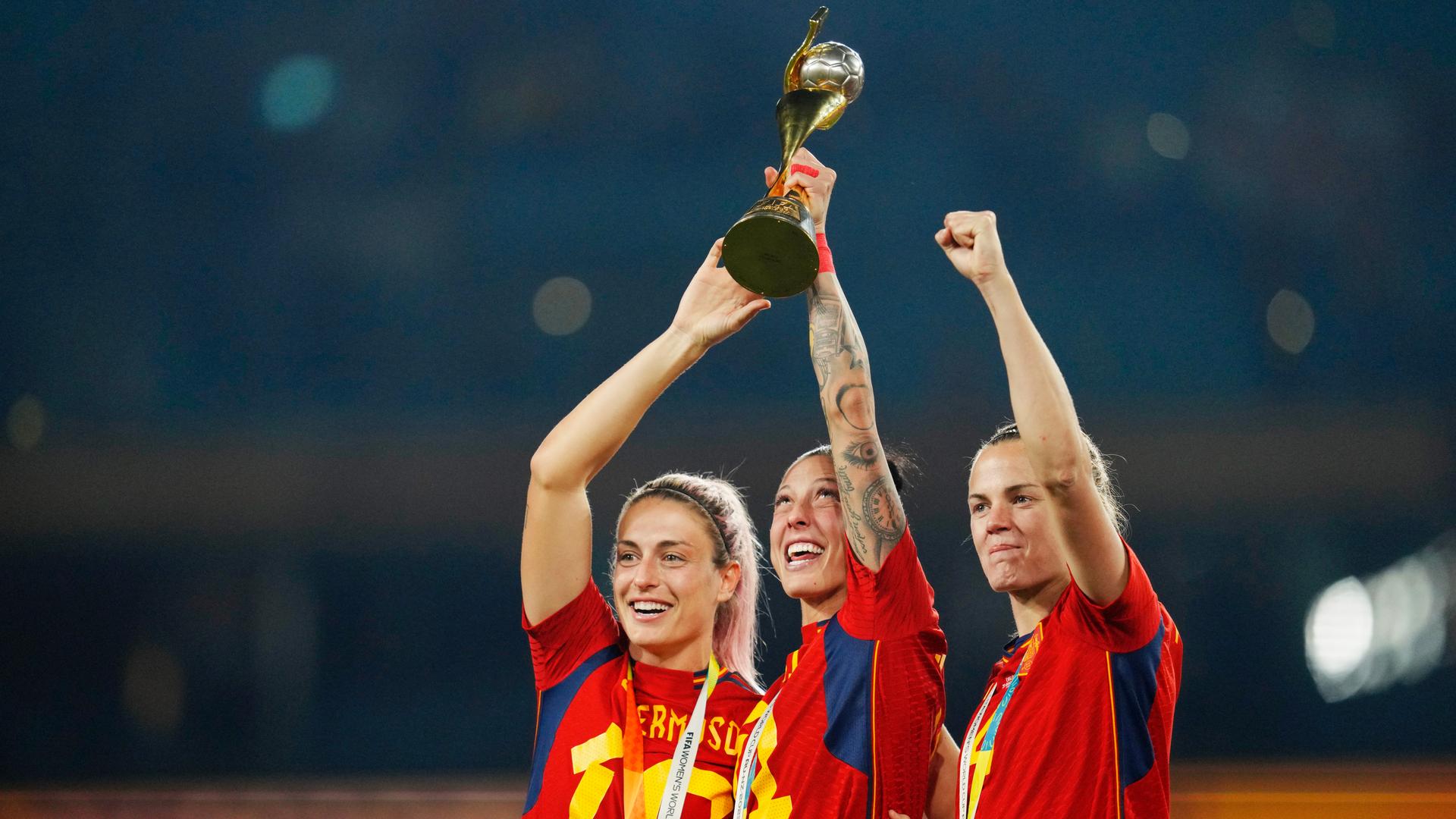 Three women in red soccer uniforms holding their hands up with a trophy