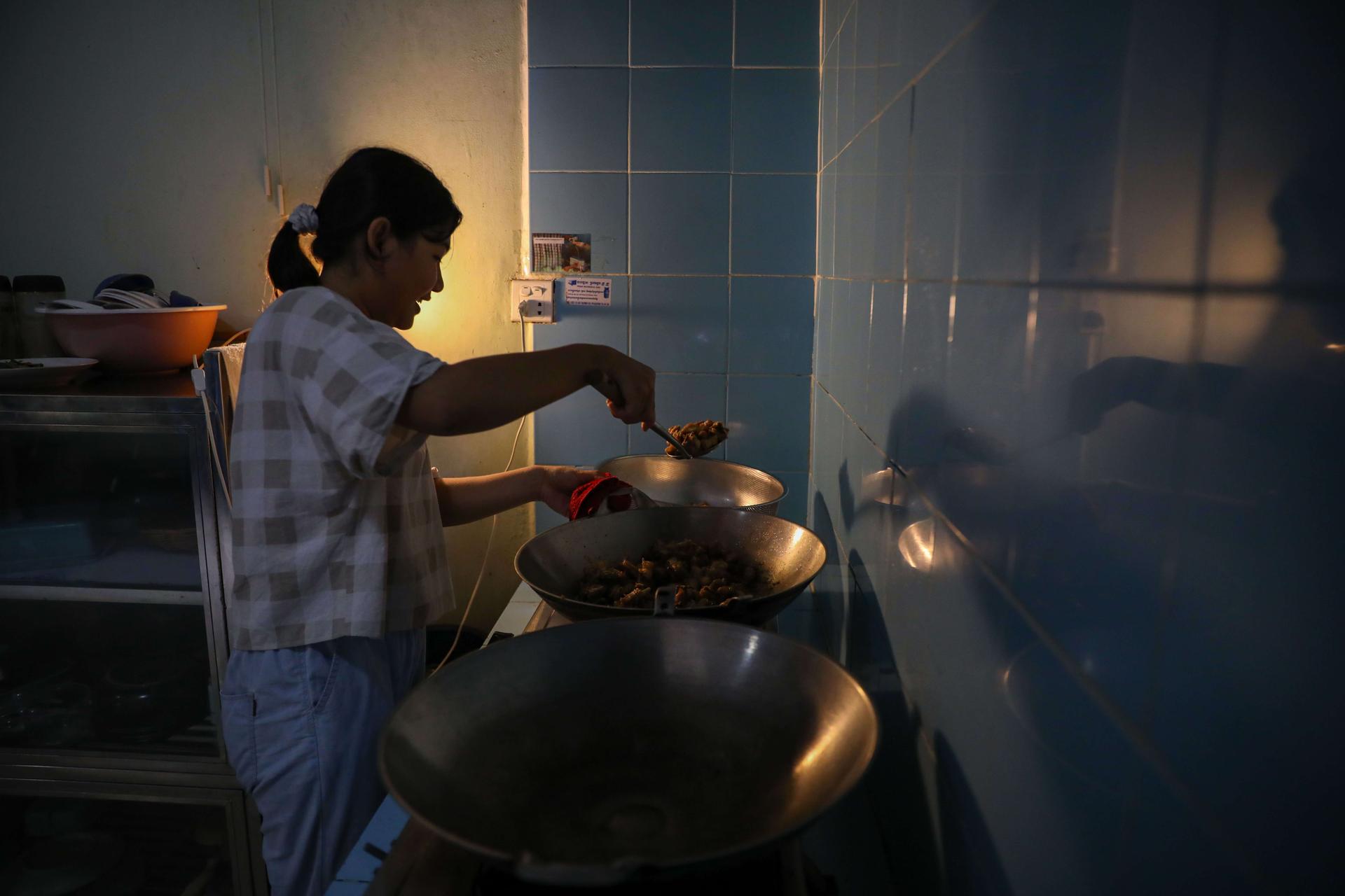 A woman seen cooking in a dimly lit kitchen, handling a wok