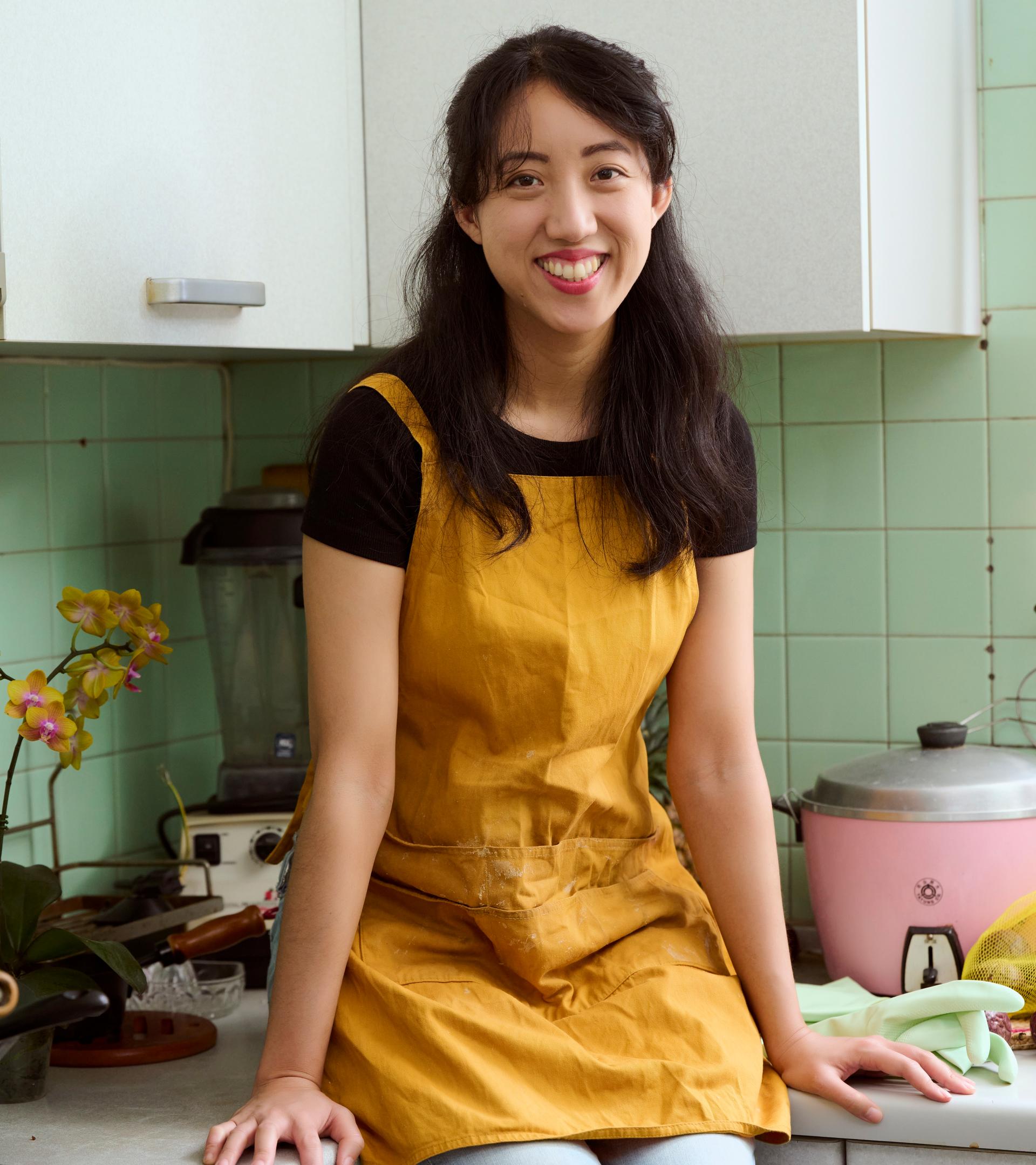 A portrait of an Taiwanese author and journalist Clarissa Wei, in a yellow apron with a kitchen backdrop.