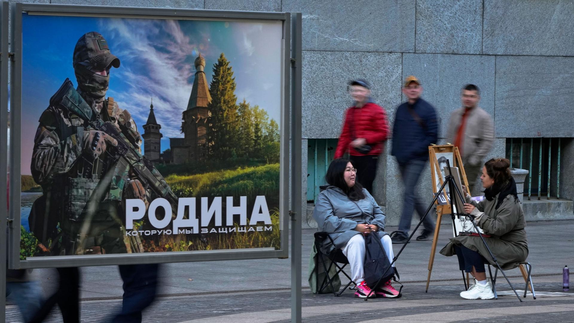 A street artist paints a portrait of a woman next to an image of a Russian serviceman and words, "The Motherland we defend," at a street exhibition of military photos, in central St. Petersburg, Russia, Friday, Oct. 6, 2023.