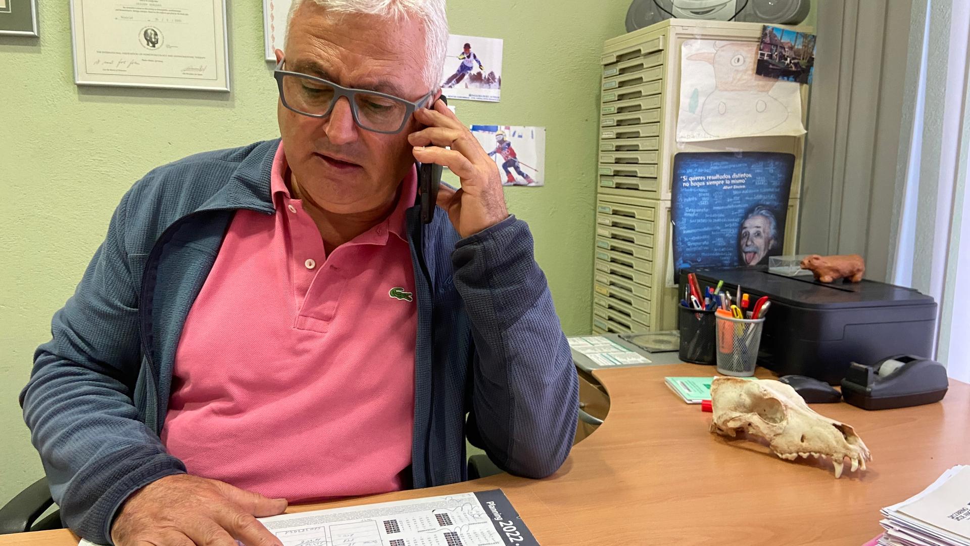 Close-up photo of a man sitting in an office on the phone. He is wearing a pink shirt and navy jacket.