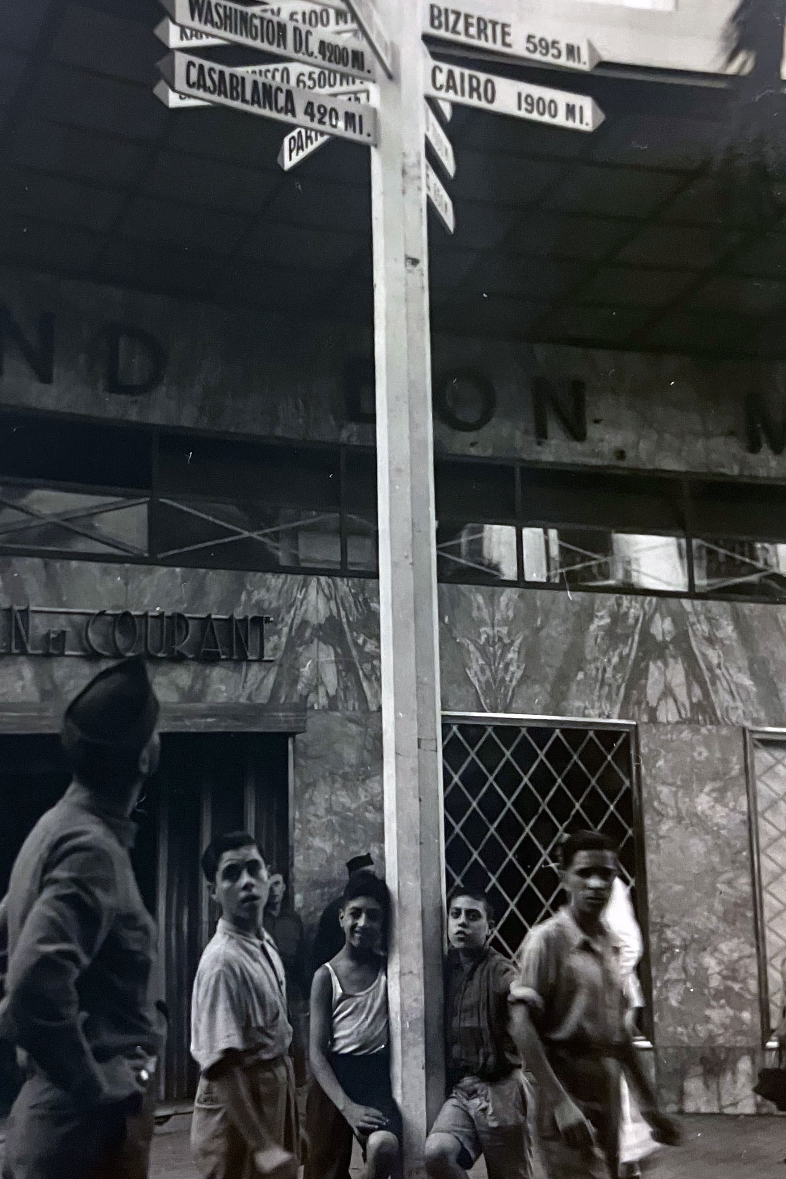 Four boys stand by a post with signs pointing to cities around the world, southern Italy, 1943.
