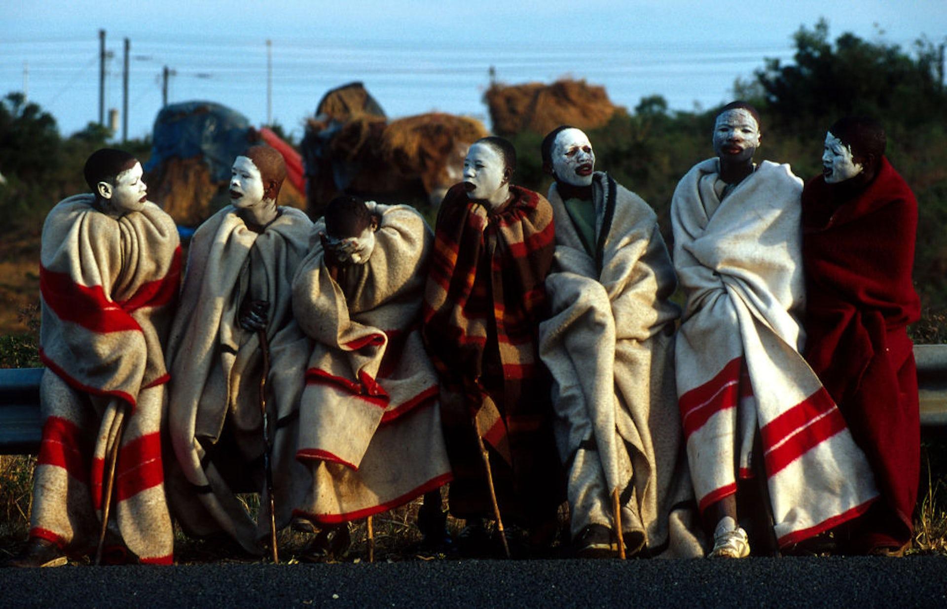 Seven boys in a row, draped in red and white blankets, their faces smeared with white clay and leaning on walking sticks.