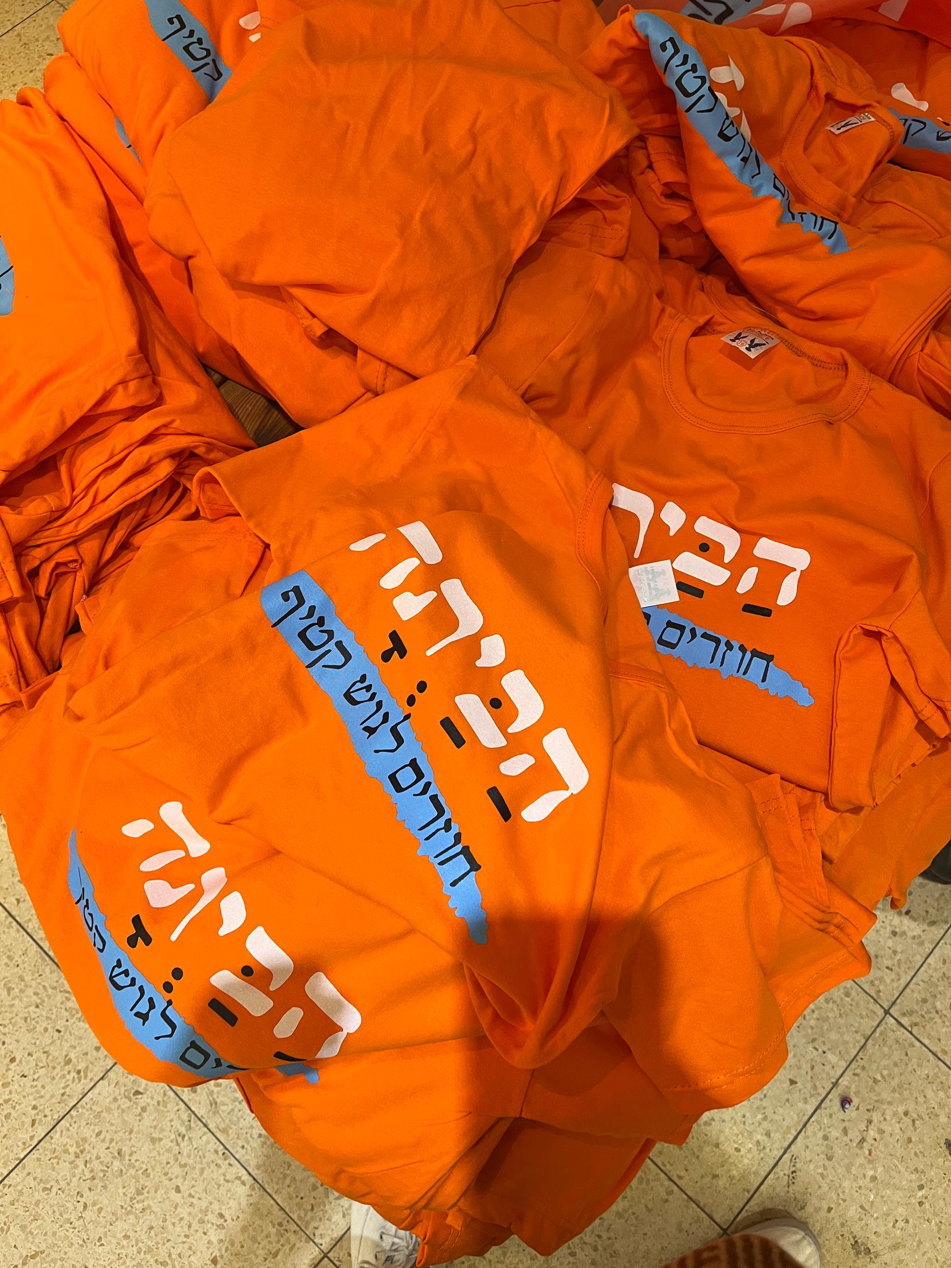 Orange T-shirts stacked in a messy pile with Israeli language written on them