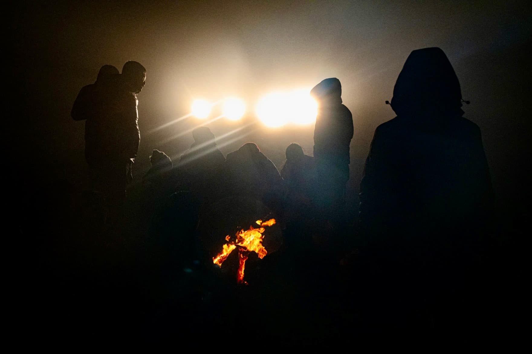 Migrants huddle by a fire as CBP trucks shine their lights on them