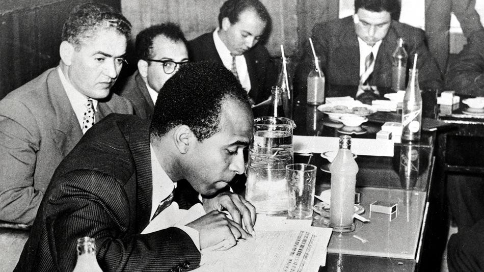 Frantz Fanon sitting at a table during a press conference 