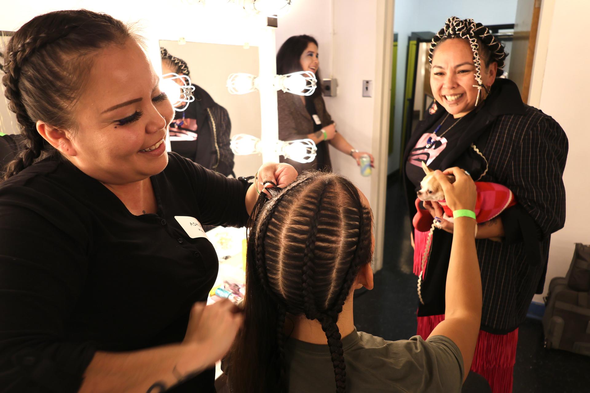 Hairstylist Ashley Jack (left) braids model Ricky-Lee Watts' (centre) hair before the Indigenous Futures streetwear fashion party.  Lauraleigh Paul (right) smiles while holding her dog.