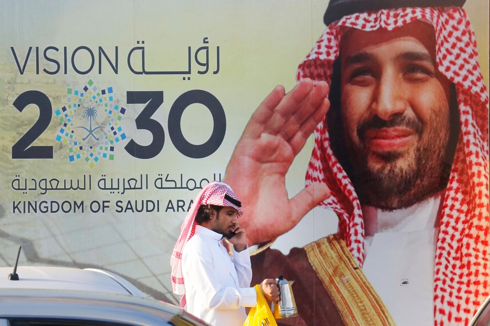 A Saudi man carries his coffee pot as he walks past a banner showing Saudi Crown Prince Mohammed bin Salman, outside a mall in Jiddah, Saudi Arabia, Feb. 5, 2020.
