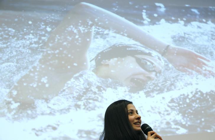 Syrian swimmer Yusra Mardini holds a microphone and addresses reporters at a news conference in Berlin. She stands before a photograph of herself projected onto a screen.