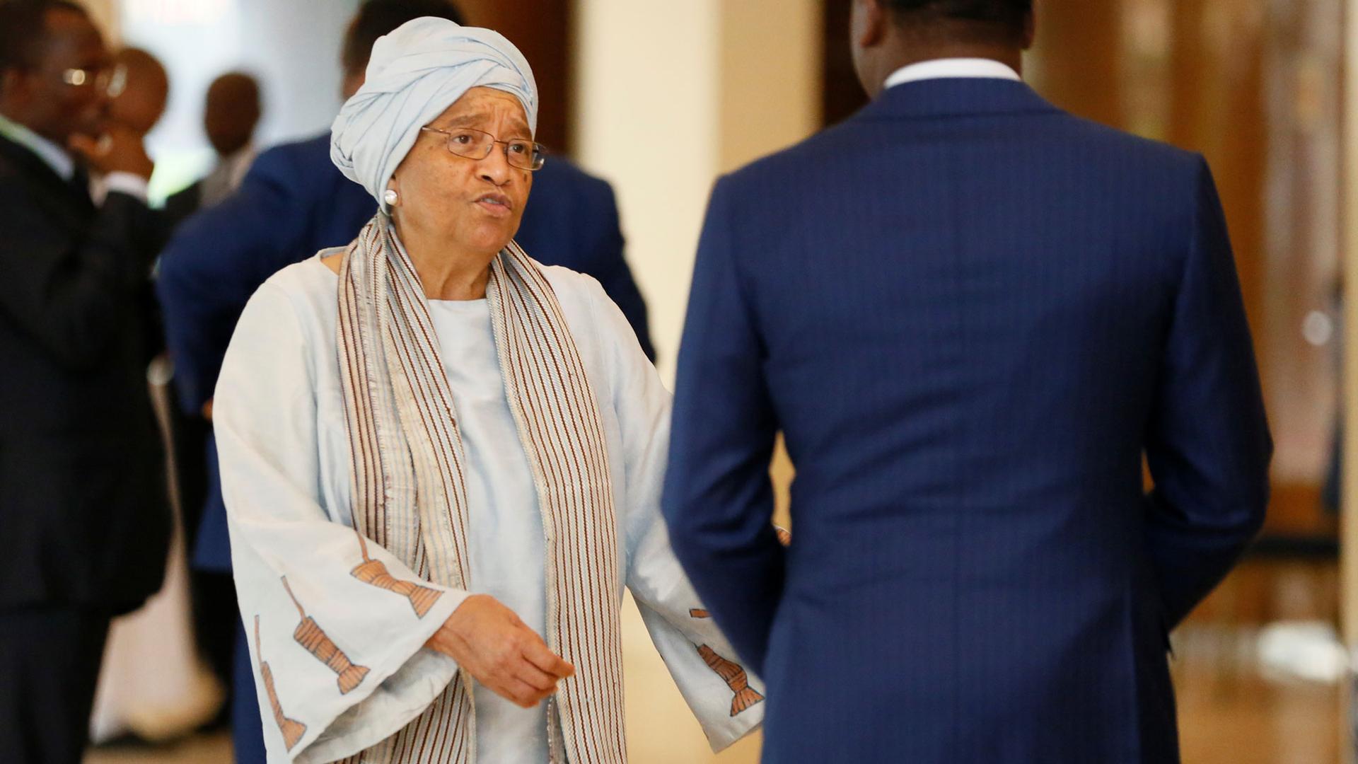 Ellen Sirleaf wears a grey headwrap and robe as she speaks to a man in a blue suit.