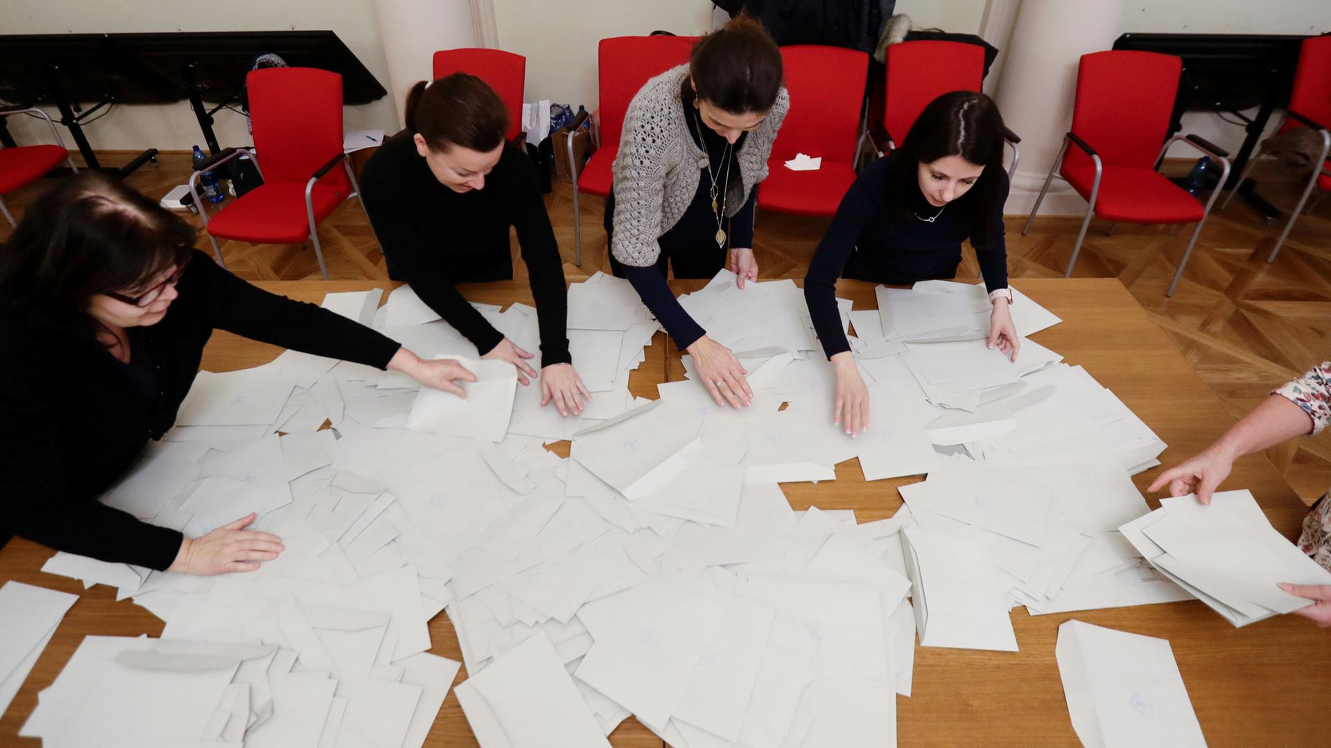 Four people sit at a table covered in white papers, which are ballots, with their hands outstretched as they sort through the pile