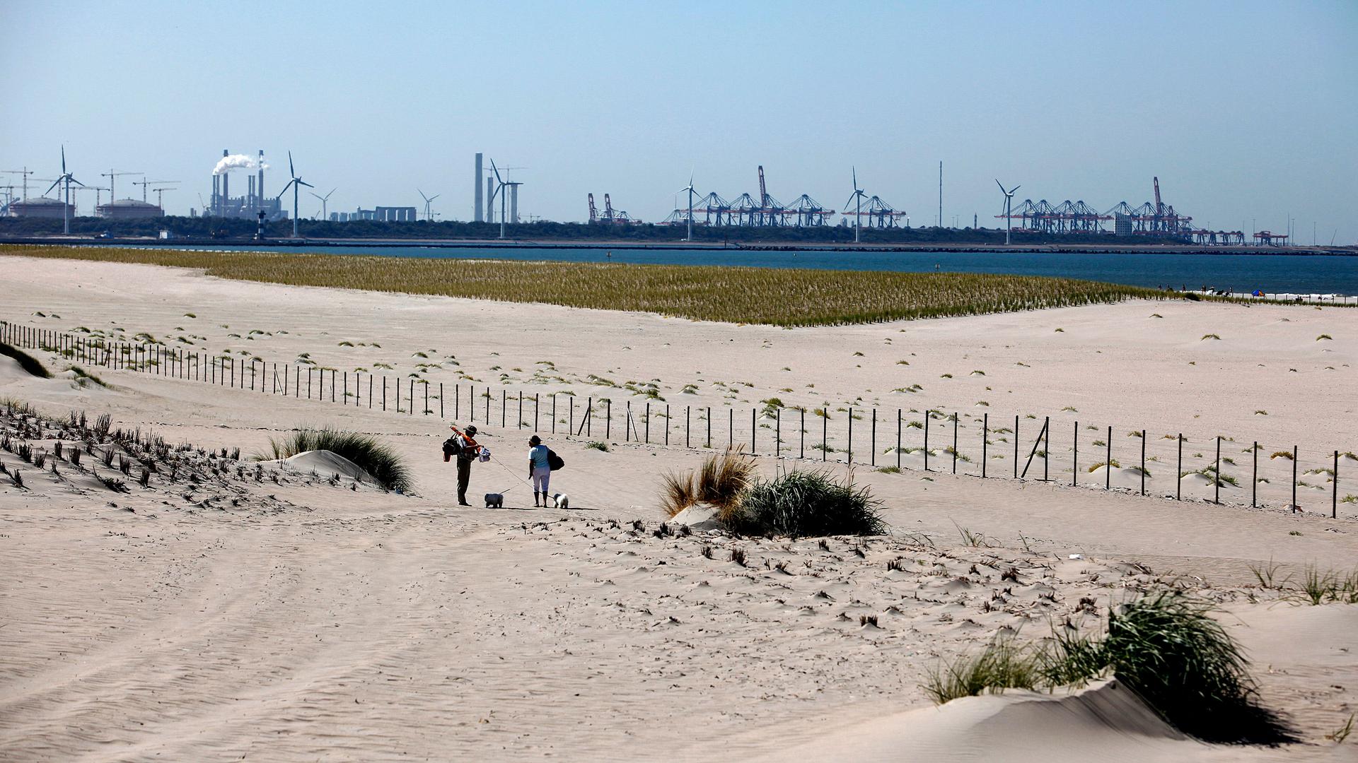 Two people walk across the beach sand. Across the water are wind turbines and other buildings